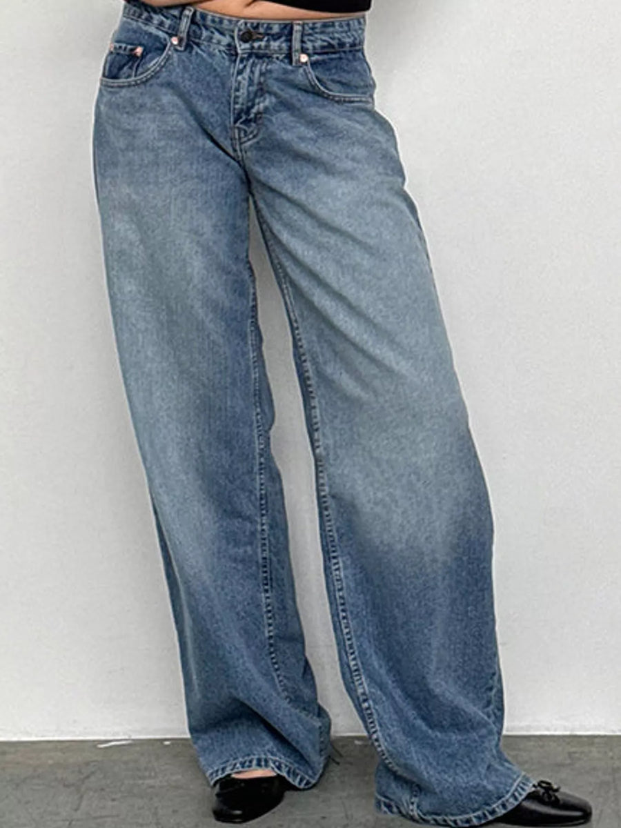 A person wearing light blue wide-leg denim jeans with black shoes, standing against a plain light gray wall.