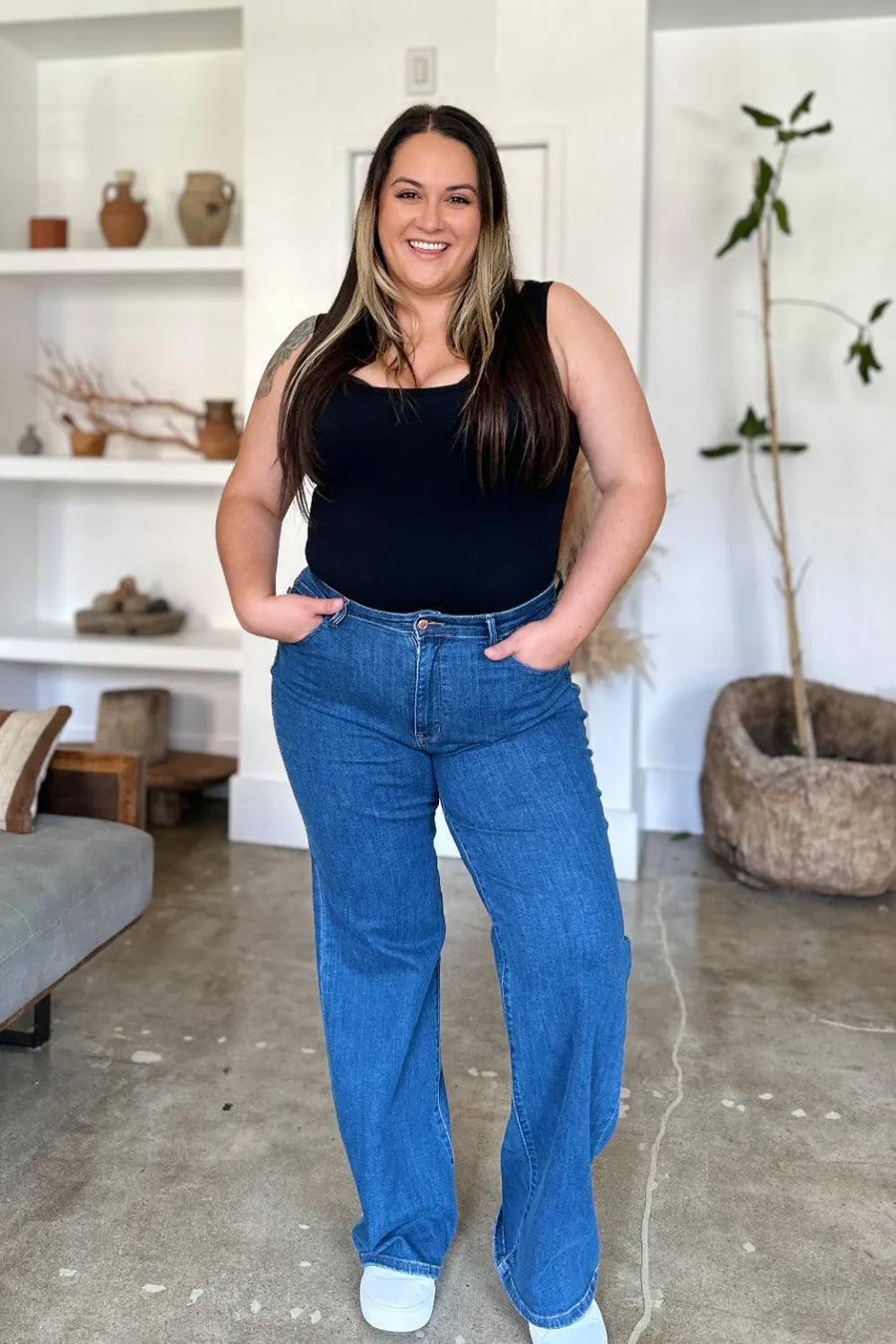 Woman wearing a black tank top and blue jeans standing in a living room.