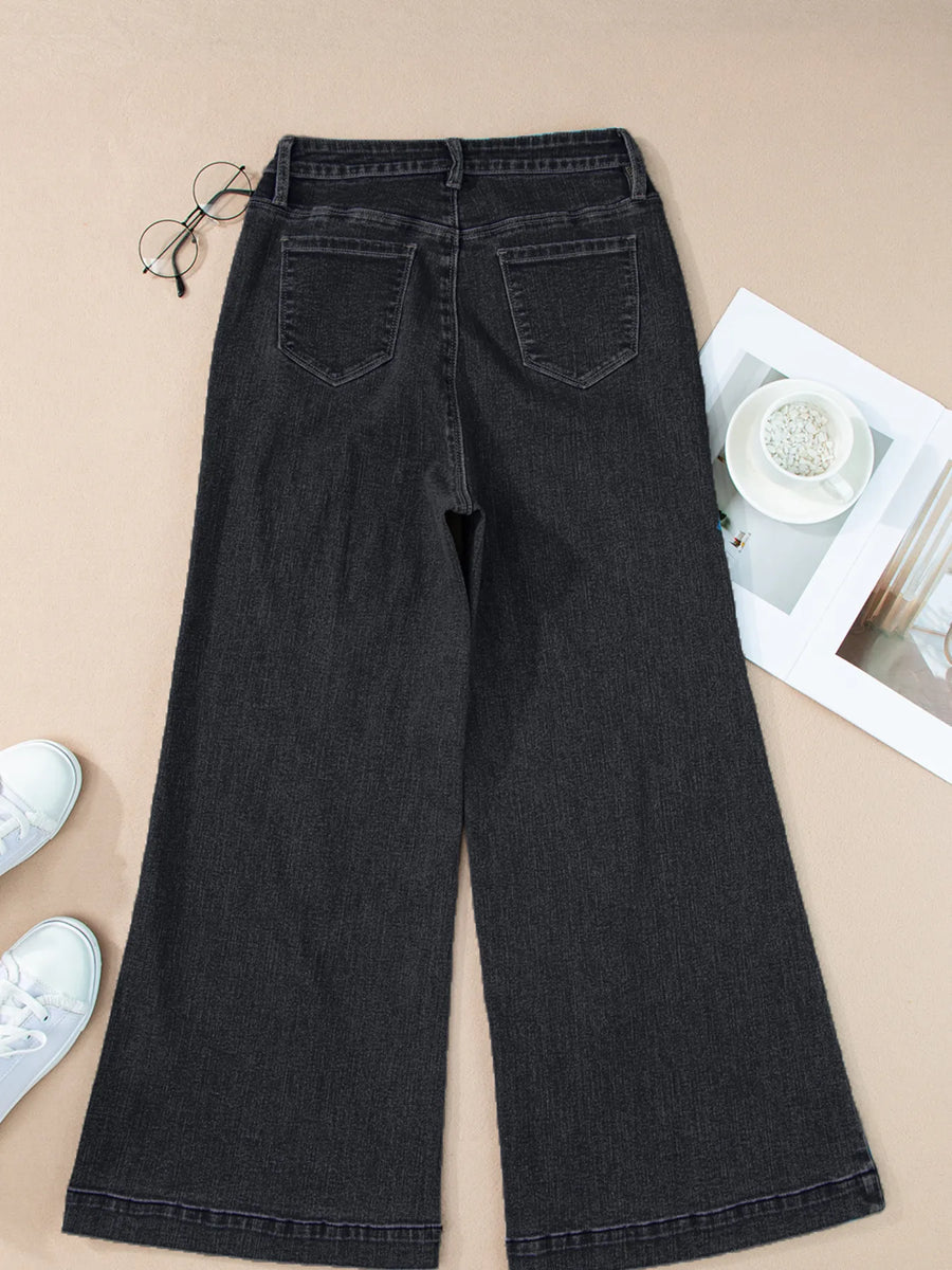 Black wide-leg jeans on a beige surface with a pair of white sneakers and a magazine.
