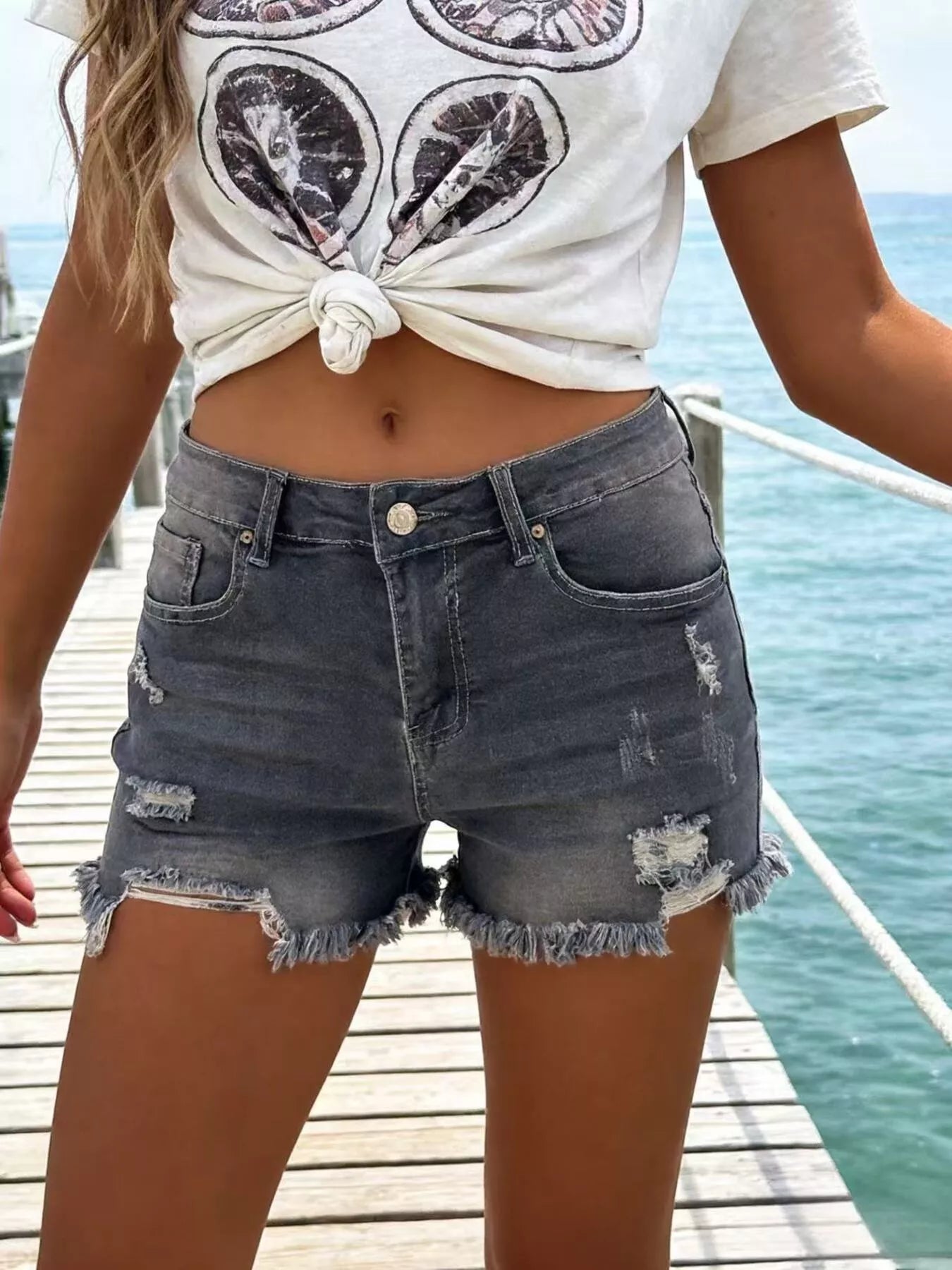 Person wearing a white crop top with black designs and denim shorts on a dock by water.