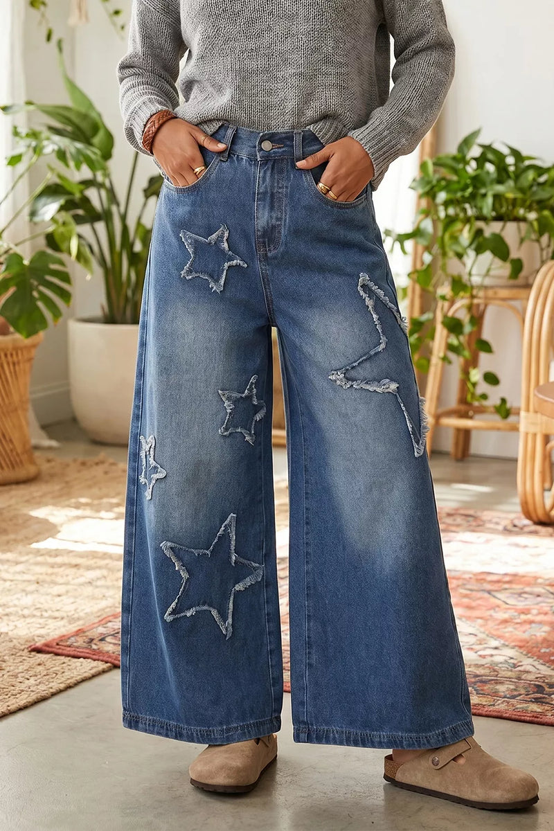A person wearing wide-leg blue denim jeans with large star-shaped patchwork details, styled with a gray knit sweater and beige slip-on shoes in a bright indoor room with plants and rugs.