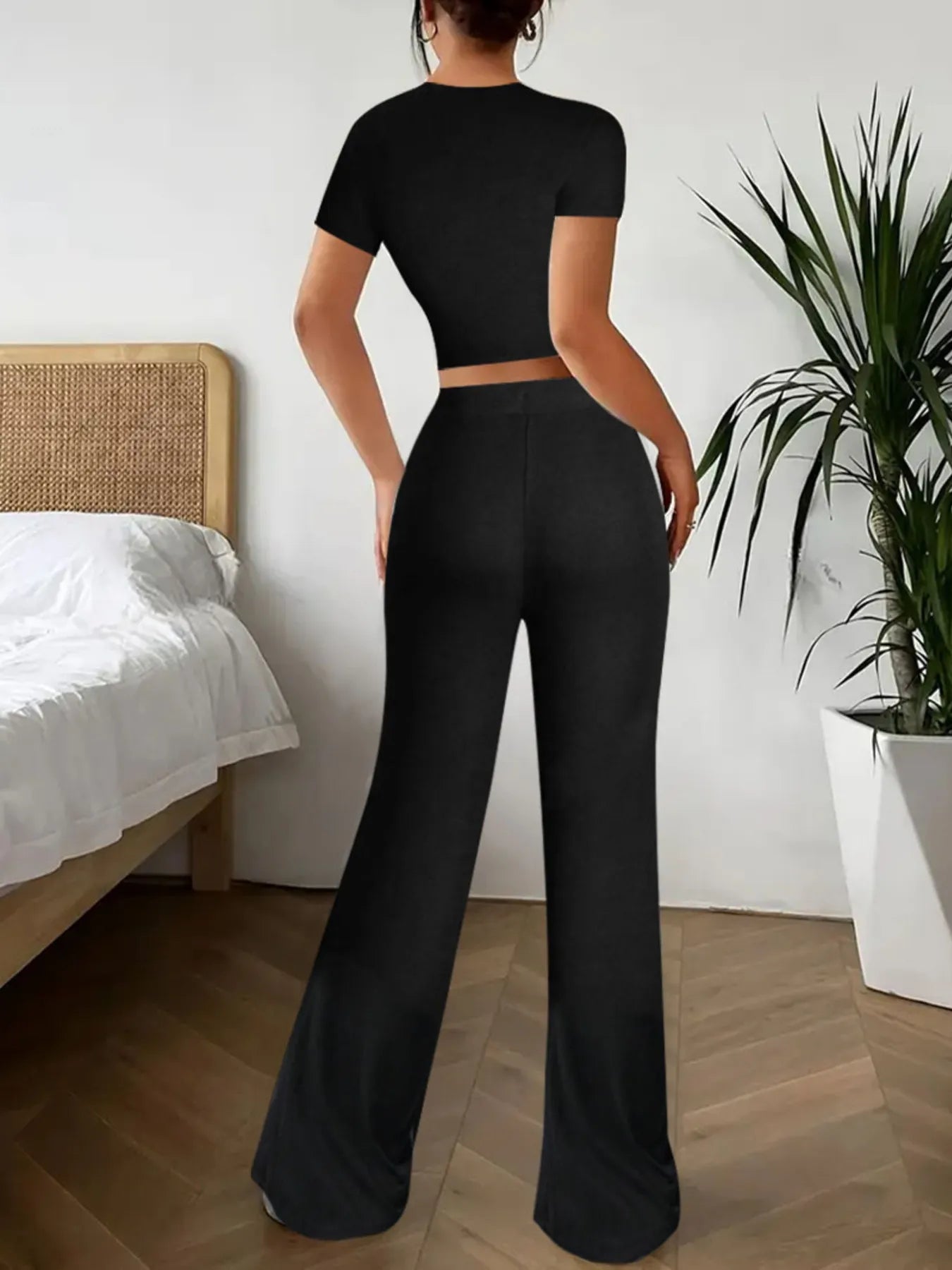 Person wearing a black outfit in a bedroom setting with a bed and plant.