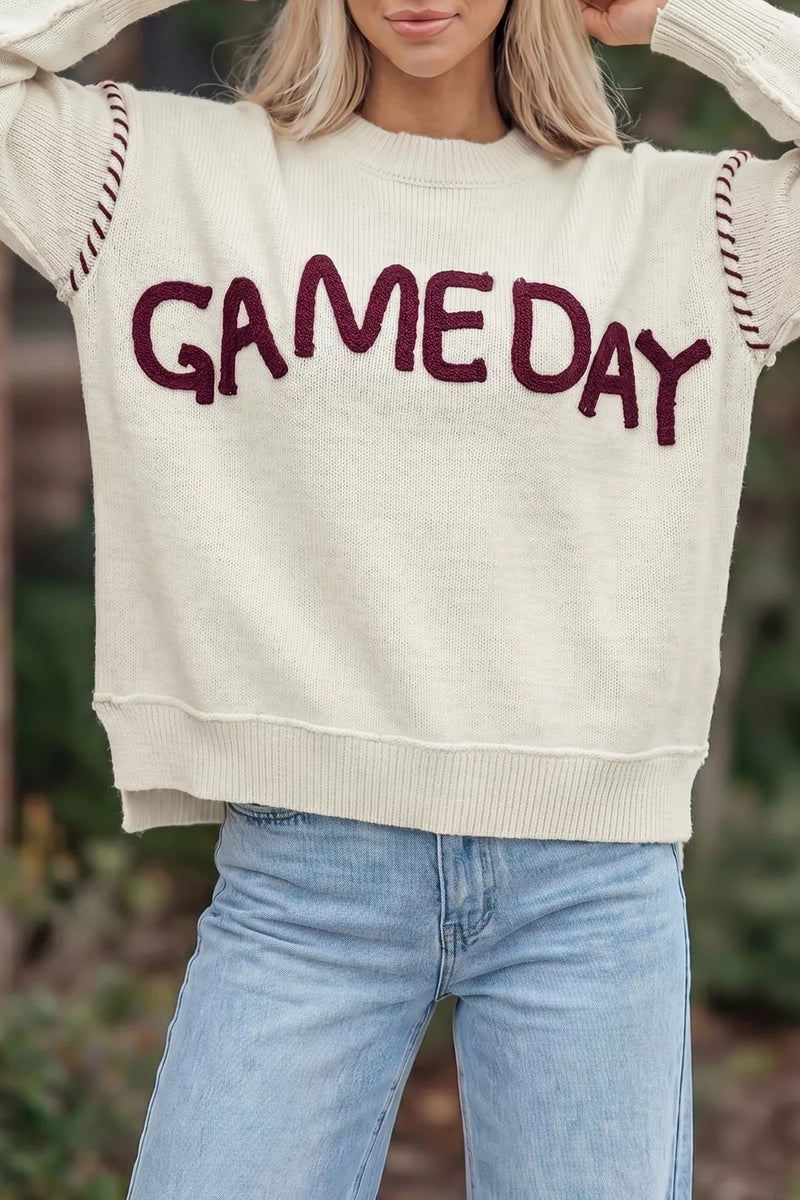A woman wearing a cream-colored knit sweater with 'GAME DAY' written across the chest in dark burgundy letters, paired with light blue jeans.