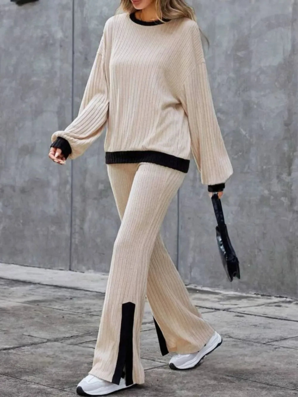 Person wearing a beige knit outfit with black accents against a gray wall.