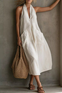 Woman wearing a white sleeveless dress holding a woven bag against a gray wall.