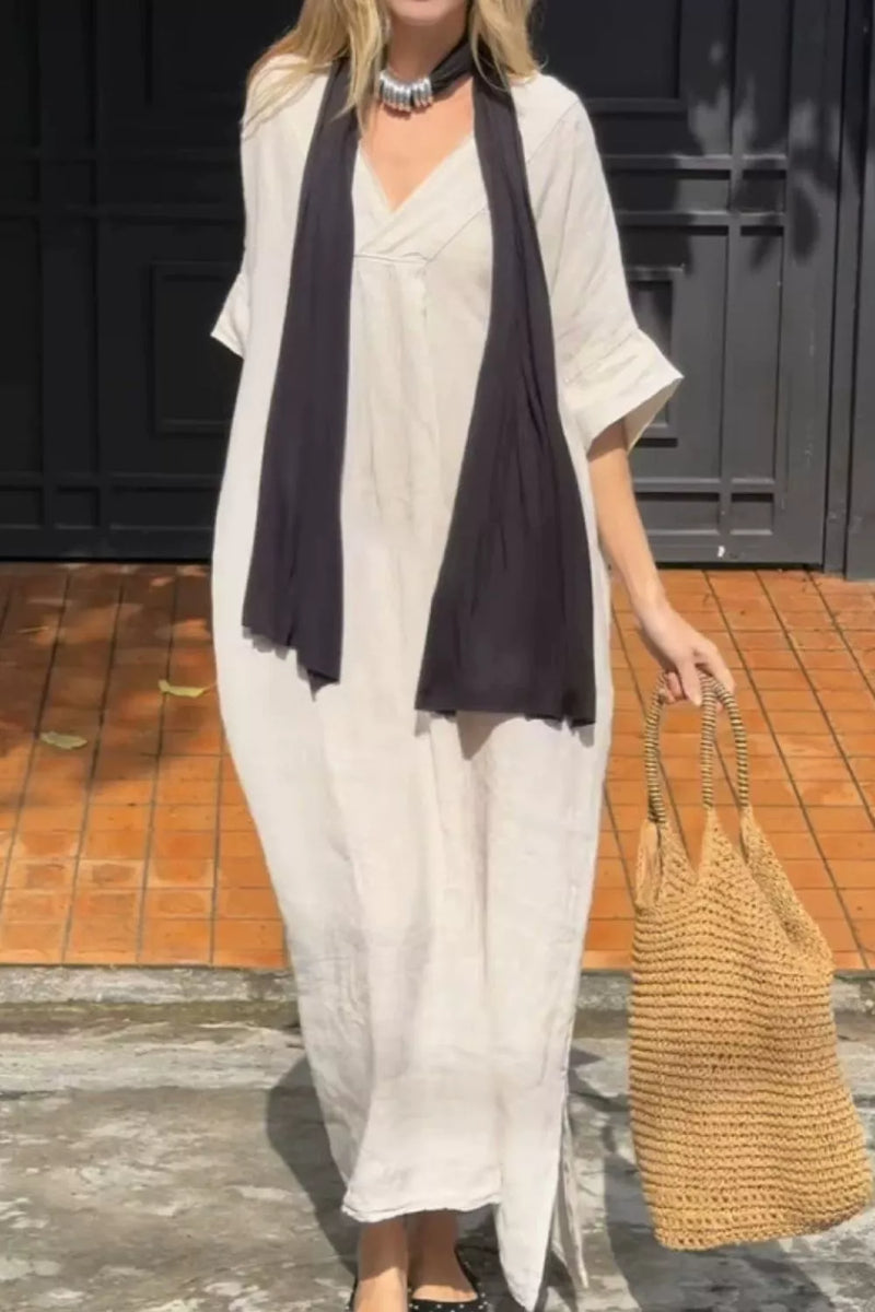 A woman wearing a loose, off-white maxi dress with wide sleeves and a dark charcoal scarf, holding a woven straw tote bag against a dark paneled door and tiled floor background.