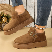 Brown suede shoes with a buckle on a beige surface, worn with blue jeans.