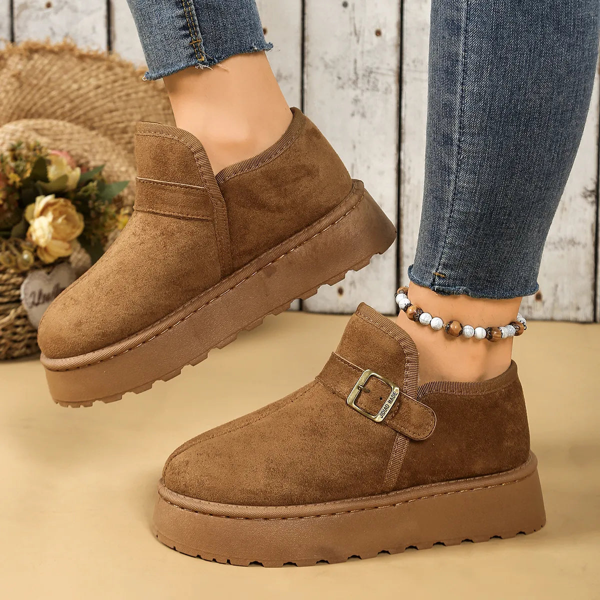 Brown suede shoes with a buckle on a beige surface, worn with blue jeans.