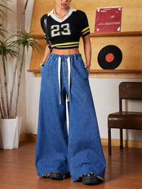 Person wearing a black and yellow sports jersey with the number 23 and blue wide-leg jeans in a room with a wooden shelf and chair.