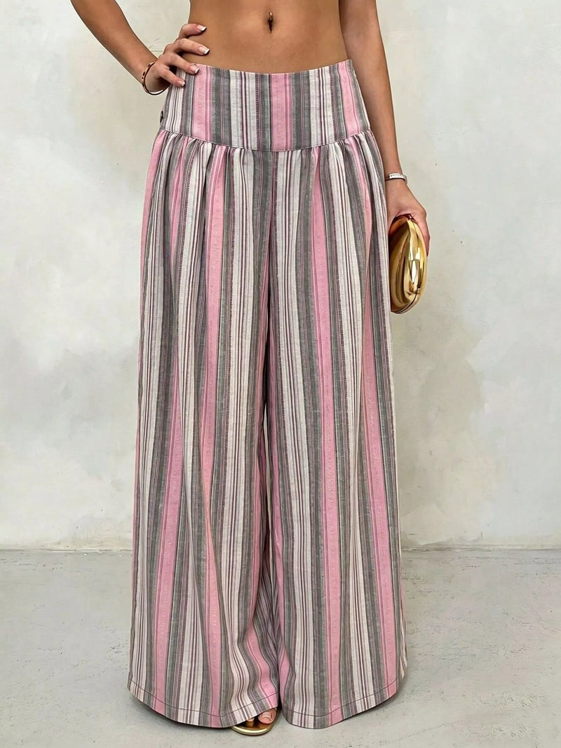 A person wearing high-waisted, wide-leg striped pants with vertical pink, gray, and white stripes, holding a small gold clutch against a textured off-white wall.