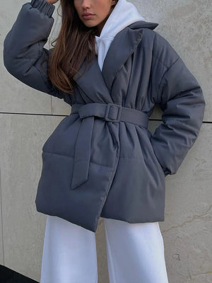 Person wearing a gray puffer coat with a belt against a neutral wall.
