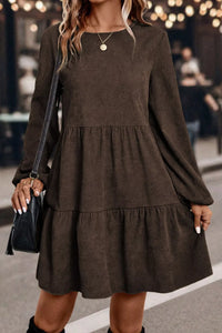 Woman wearing a brown dress with long sleeves and layered skirt, holding a black handbag.