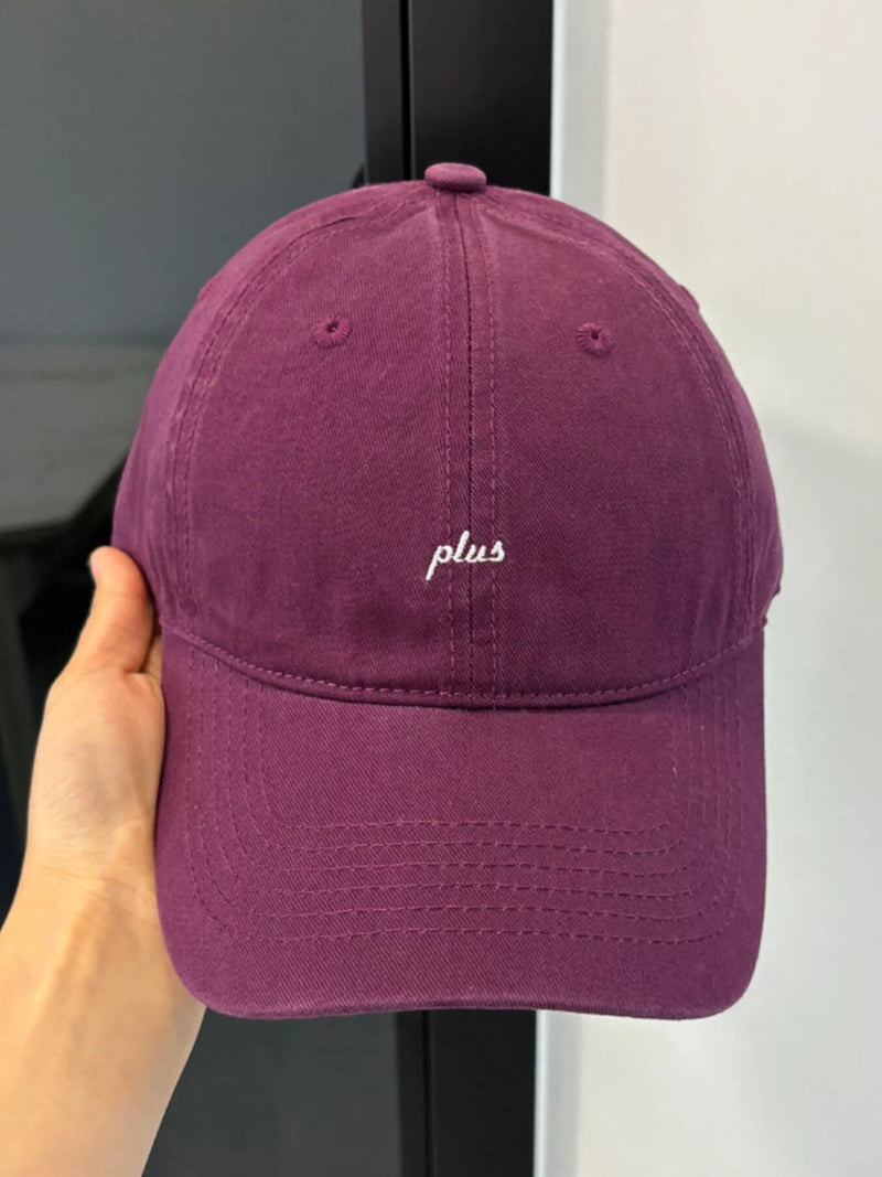 Purple cap with 'plus' embroidery held by a hand against a neutral background