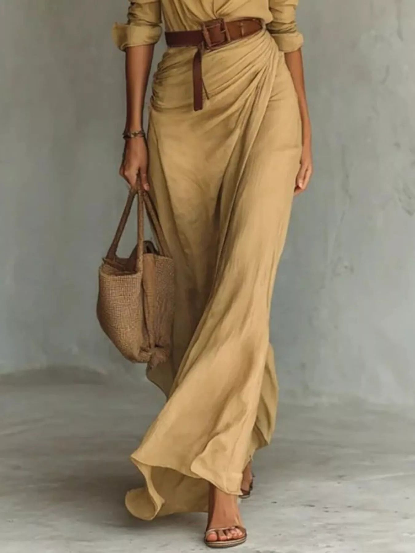 Woman wearing a long beige dress with a belt, holding a woven bag against a neutral background