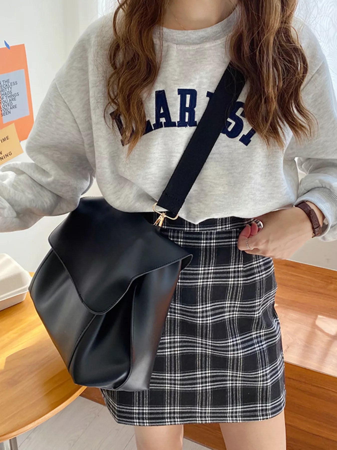 Person wearing a gray sweatshirt with 'LARSS' text and a plaid skirt, holding a black bag.