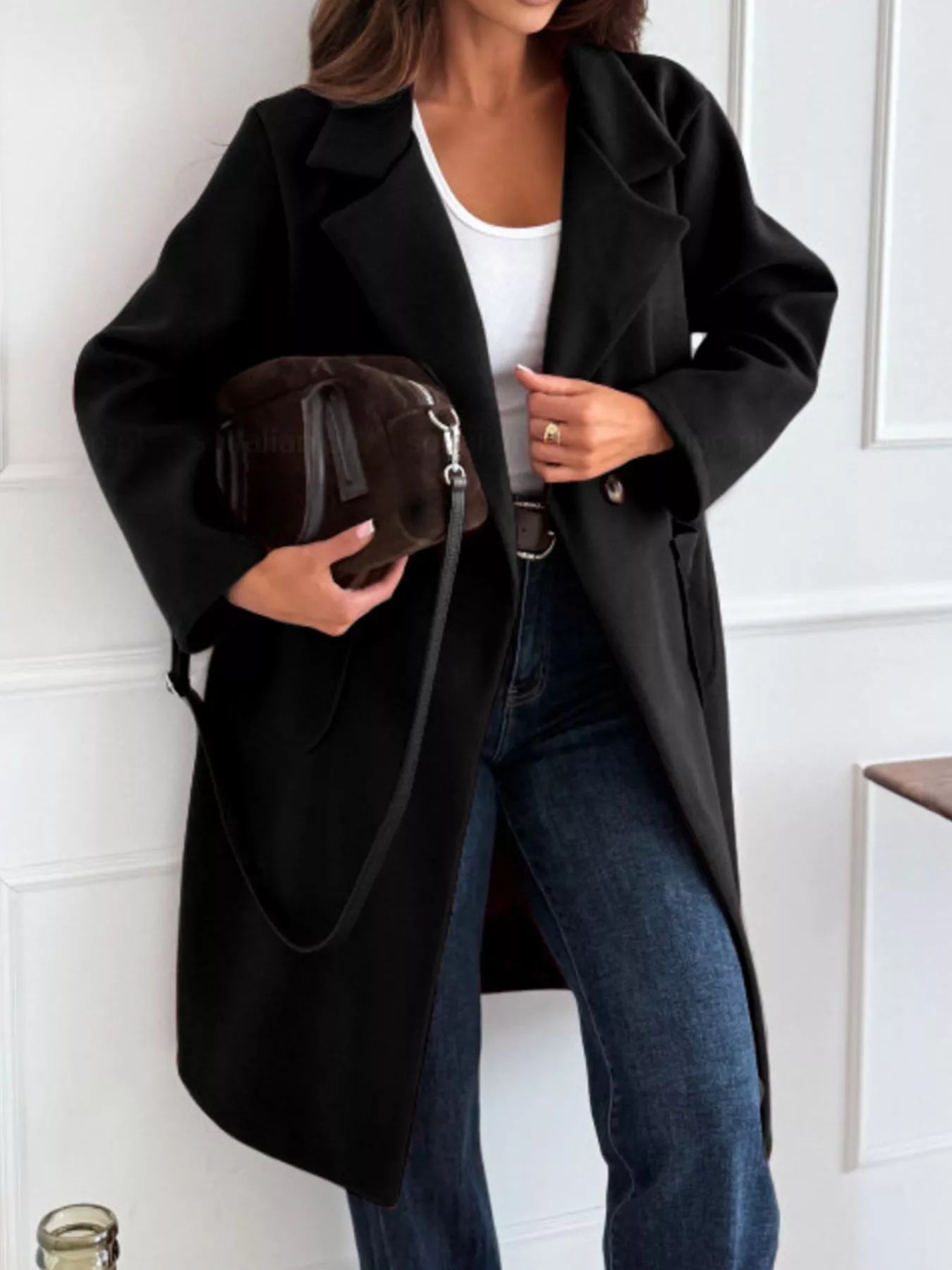 Person wearing a black coat holding a brown handbag against a white wall.