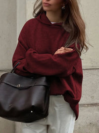 Person wearing a burgundy sweater holding a black bag against a neutral background