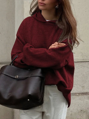Person wearing a burgundy sweater holding a black bag against a neutral background