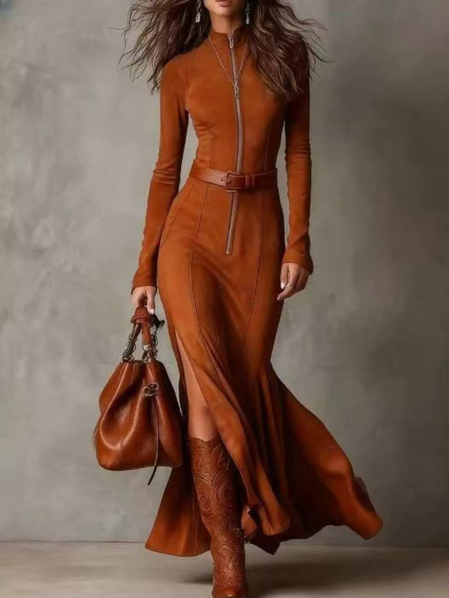 Woman wearing a long brown dress with a belt and holding a matching handbag against a gray background