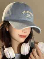 Person wearing a denim cap with 'Brooklyn' embroidery and holding headphones, on a neutral background