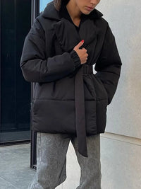 Person wearing a black puffer coat with a belt, standing against a light-colored wall.