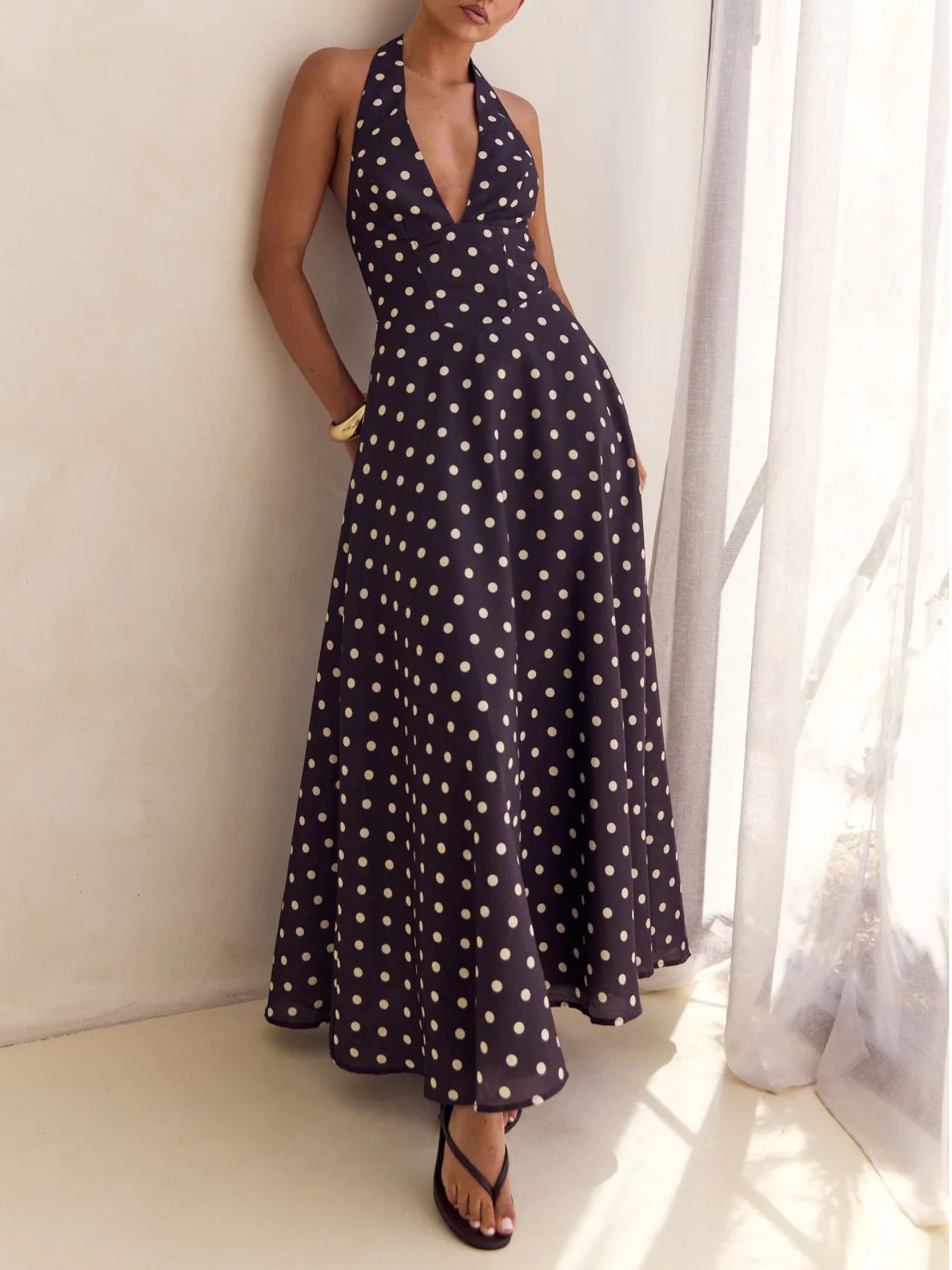 Woman wearing a black polka dot dress standing against a white wall with a curtain.