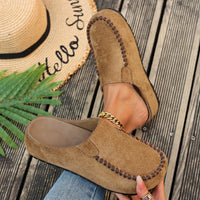 Brown suede loafers worn with a straw hat and palm leaf on a wooden surface