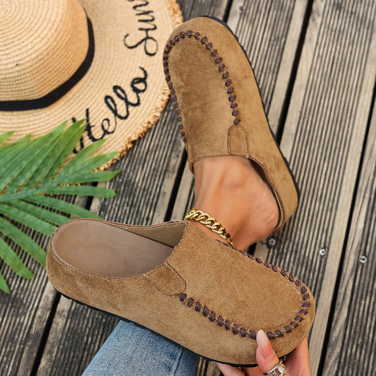 Brown suede loafers worn with a straw hat and palm leaf on a wooden surface
