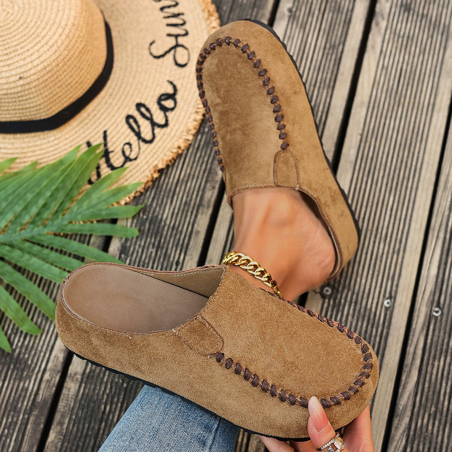 Brown suede loafers worn with a straw hat and palm leaf on a wooden surface