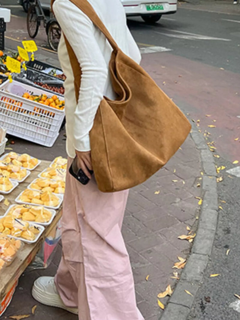 A person wearing a white long-sleeve top and light pink pants carries a large tan suede shoulder bag while standing on a city sidewalk next to a fruit stand.