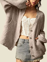 A woman wearing a chunky, oversized light taupe knit cardigan over a white lace-trim crop top and a denim mini skirt, set against a solid light beige background.