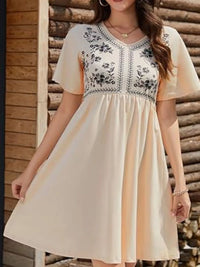 Woman wearing a beige dress with blue embroidery against a wooden background