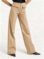 A model is wearing a pair of beige wide-leg trousers with front flap pockets and a zippered waist, paired with black pointed shoes on a light gray studio background.