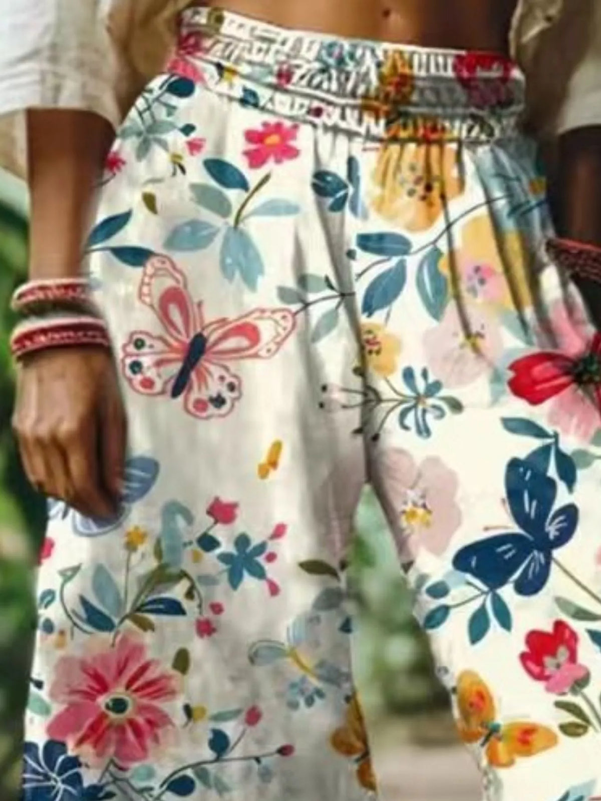 A close-up of a person wearing loose, high-waisted white pants with a colorful floral and butterfly print. The print features pink, blue, yellow, and green motifs on a white base, with a patterned elastic waistband.