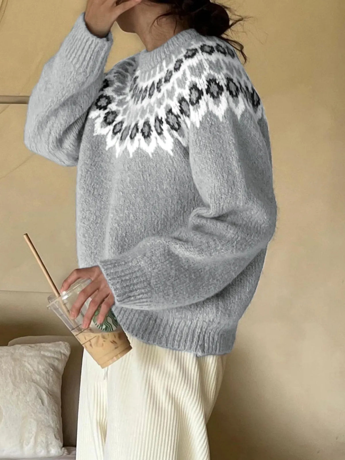 Person wearing a gray sweater with a decorative pattern, holding a drink and a book.