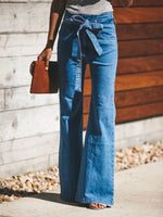 Person wearing blue flared jeans holding a brown leather bag against a wooden and stone wall.