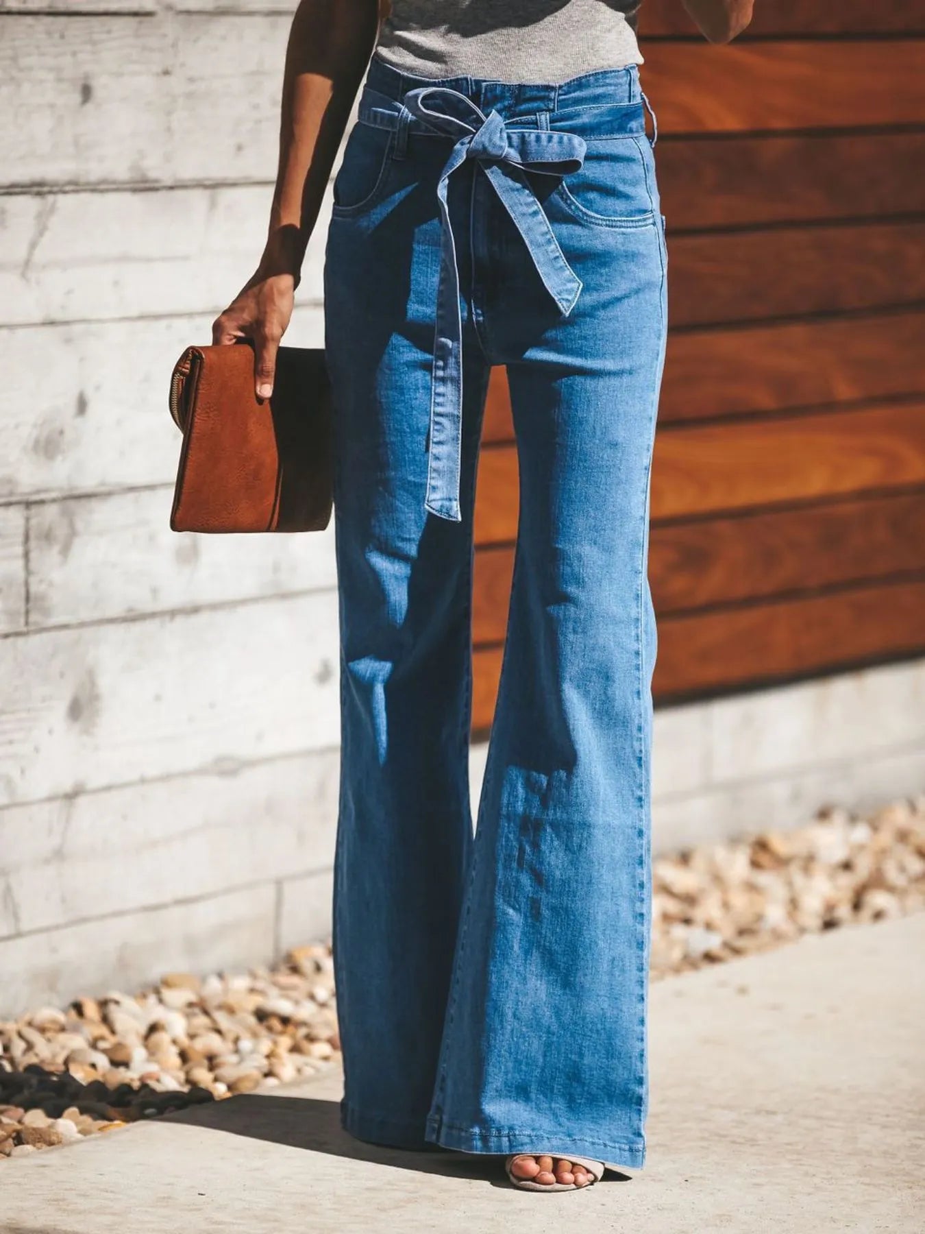 Person wearing blue flared jeans holding a brown leather bag against a wooden and stone wall.