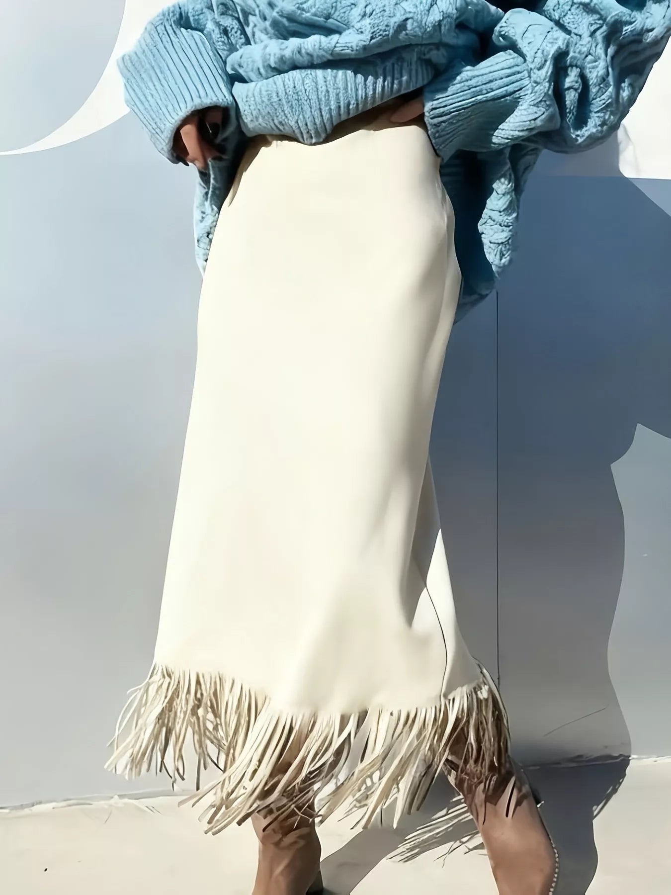 Person wearing a blue knitted sweater and white pants with fringe, standing against a light gray background.