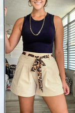 Woman wearing a navy top and beige shorts with a leopard print belt in a room.