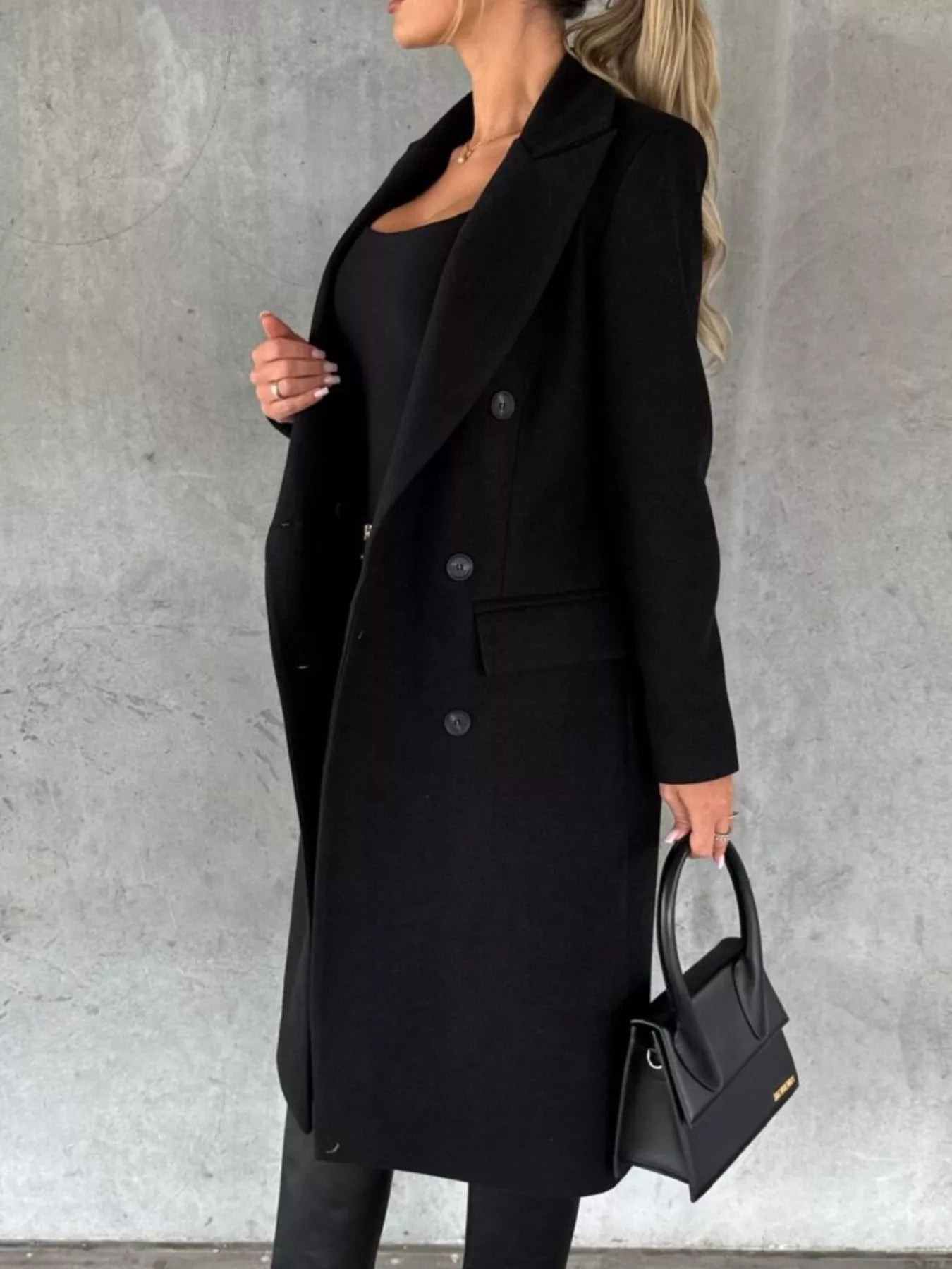 Person wearing a black coat holding a black handbag against a gray background