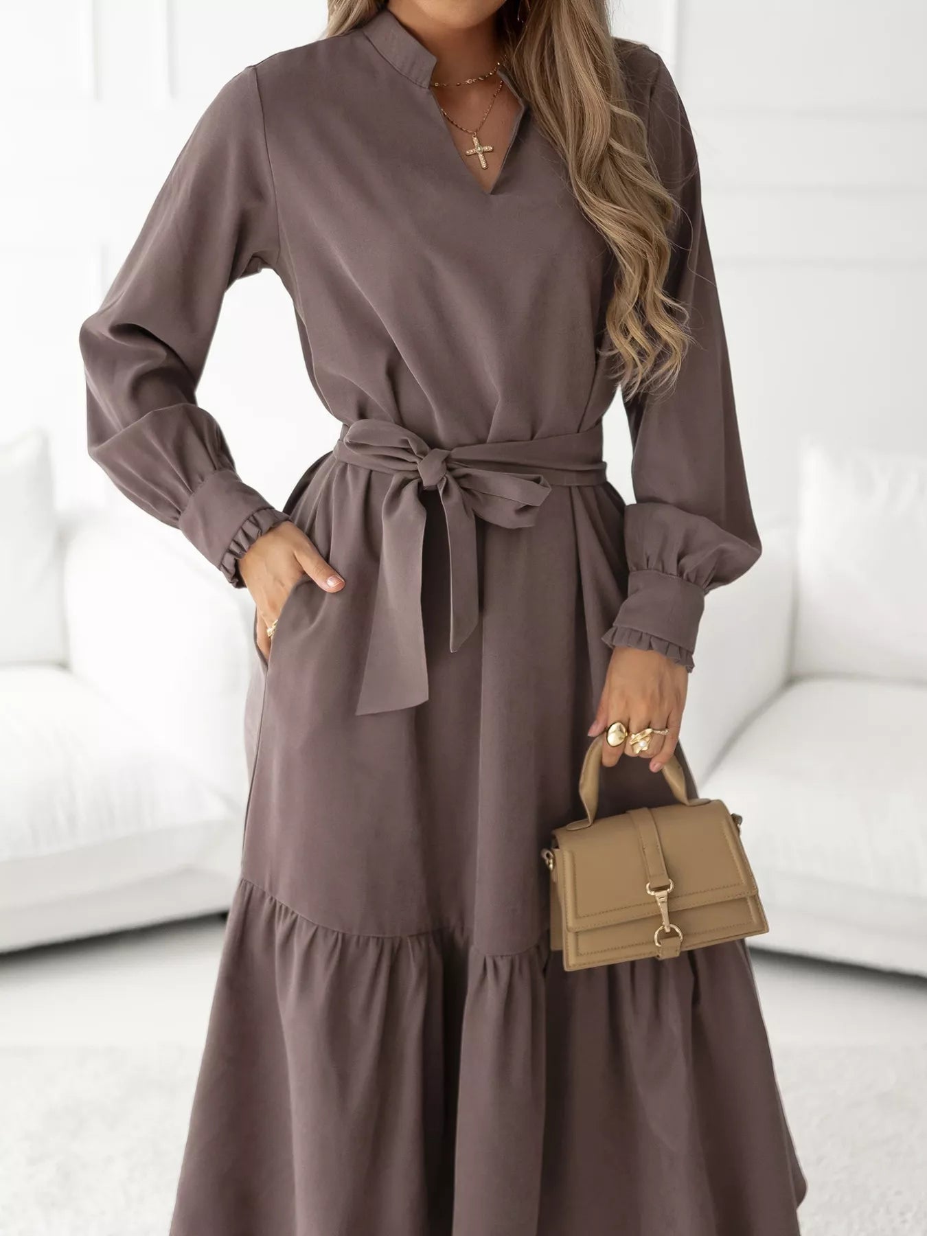 Woman wearing a long brown dress with a belt, holding a small handbag indoors.