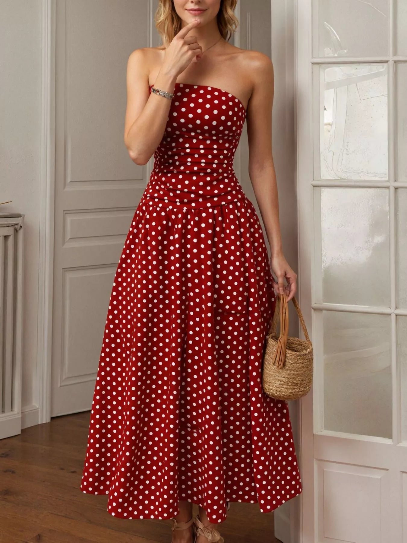 Woman wearing a red strapless dress with white polka dots, holding a woven bag indoors.