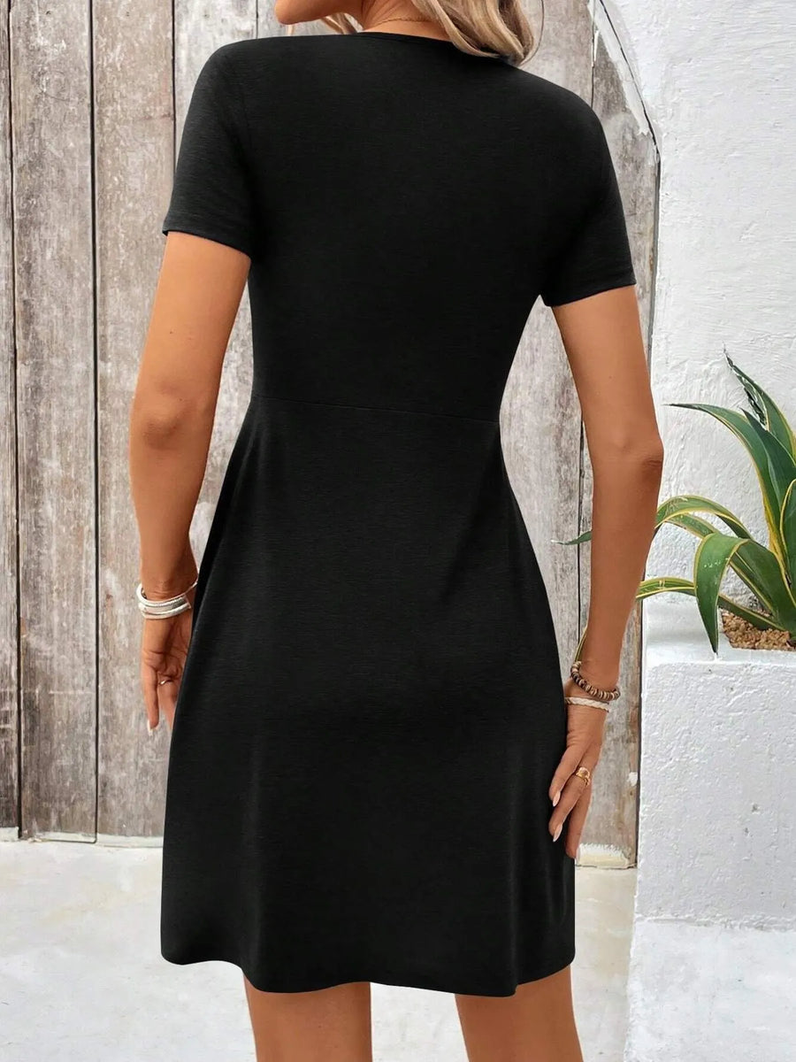 Person wearing a black dress standing against a wooden and white wall with a plant in the background.
