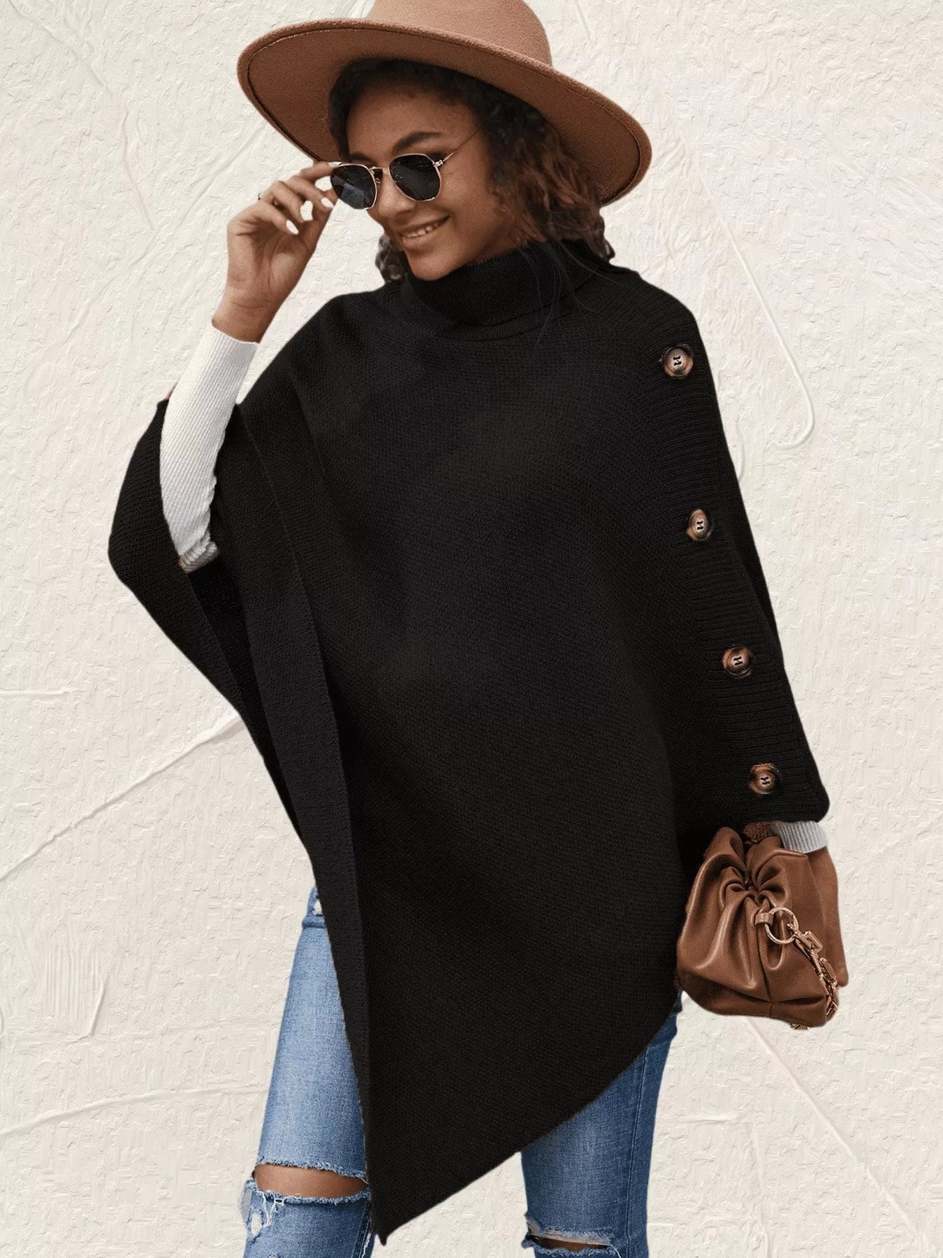 Woman wearing a black poncho with buttons, blue jeans, and a brown hat on a light background