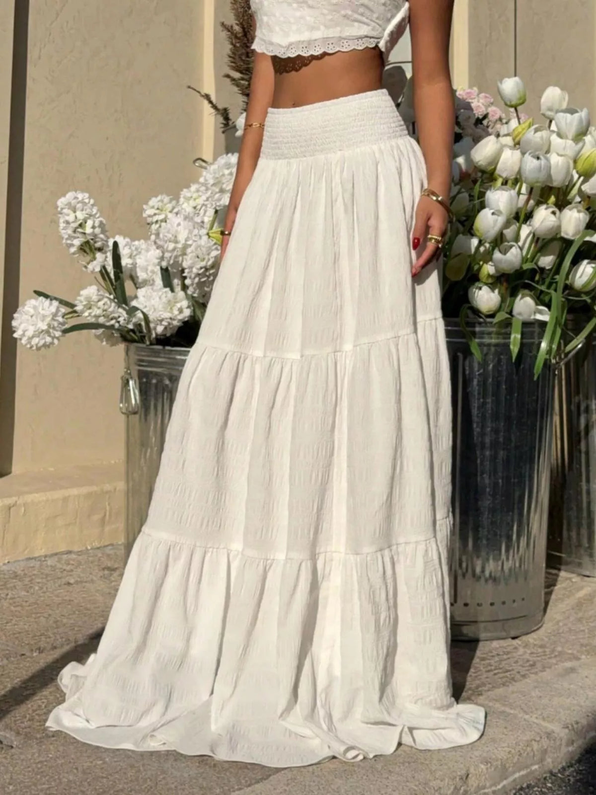 Person wearing a white dress standing between two flower pots with white flowers.