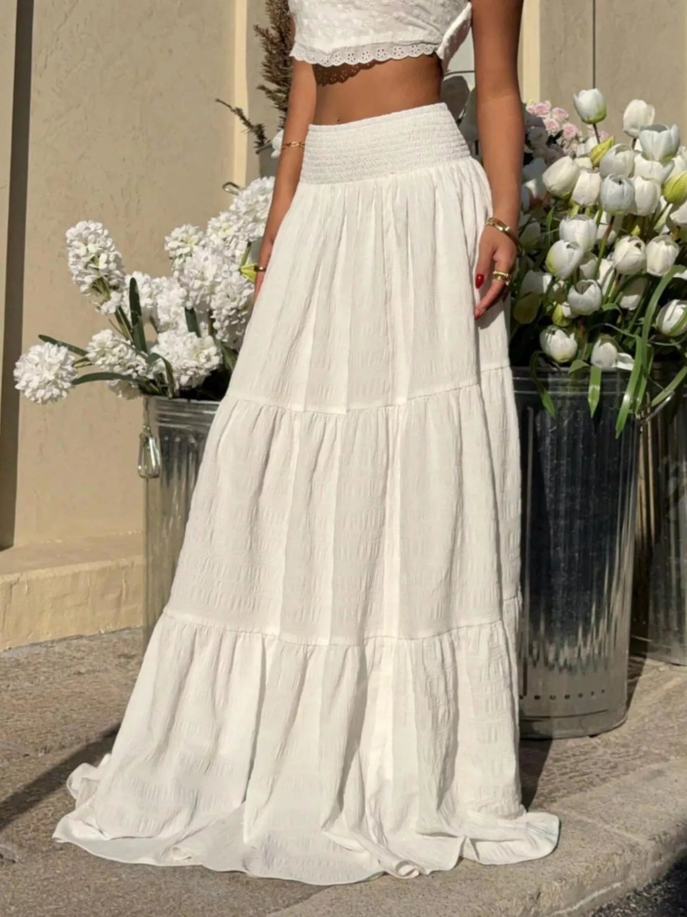 Person wearing a white dress standing between two flower pots with white flowers.