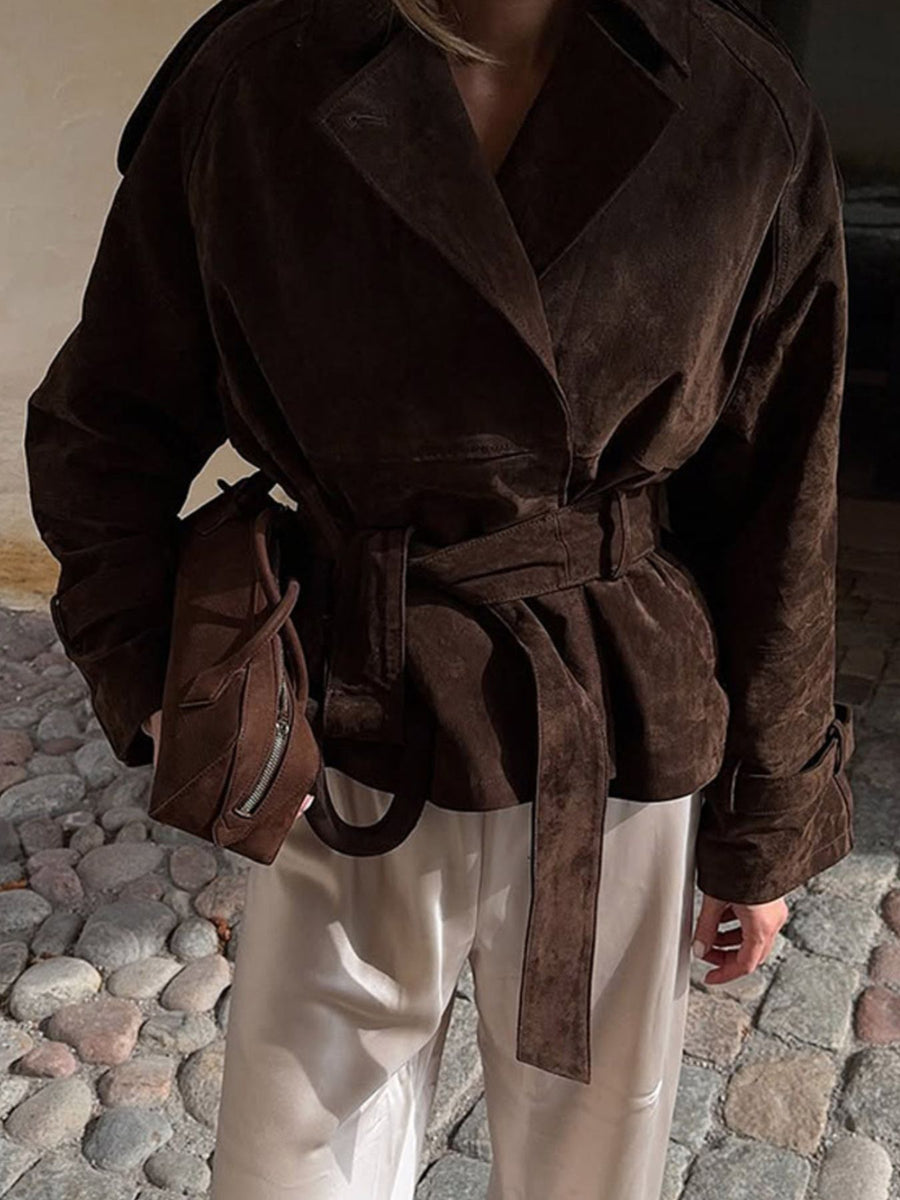 Person wearing a brown suede coat with a belt, standing on a stone pavement.