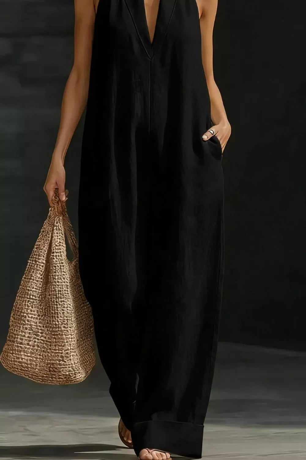 Person wearing a black sleeveless dress holding a woven bag against a dark background