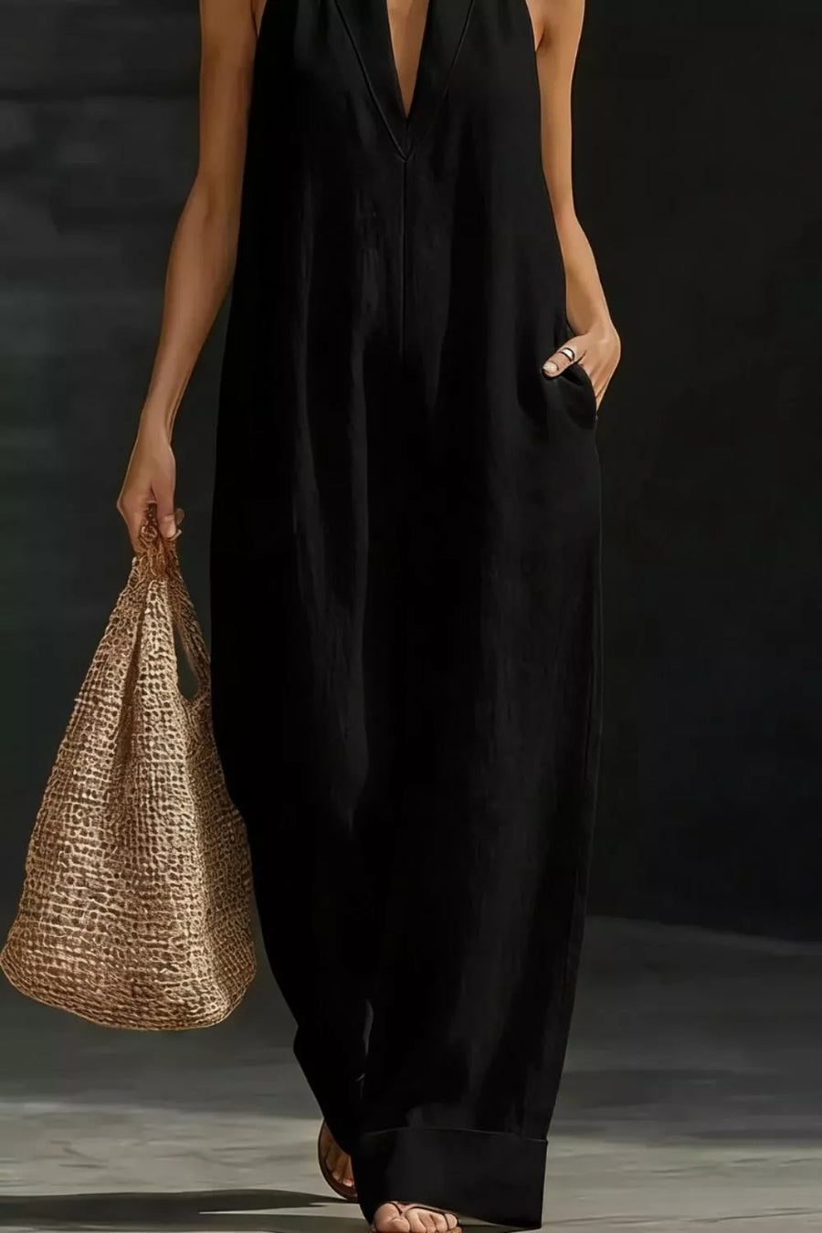 Person wearing a black sleeveless dress holding a woven bag against a dark background