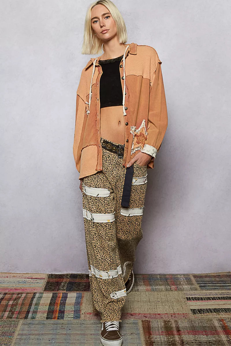 Person wearing a tan jacket and patterned pants standing against a plain background